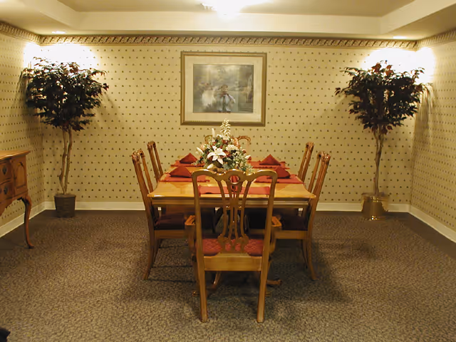 A formal dining room with a wooden dining table set for six people. The table has a floral centerpiece and red napkins on each place setting. The room features patterned wallpaper, two potted artificial trees on either side, a framed painting on the wall, and carpeted flooring.