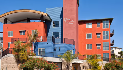 Exterior view of a modern multi-story senior living facility with red and blue walls, balconies, palm trees, and landscaping under a clear blue sky. The building has a sign that reads 'Plaza Village Senior Living'.