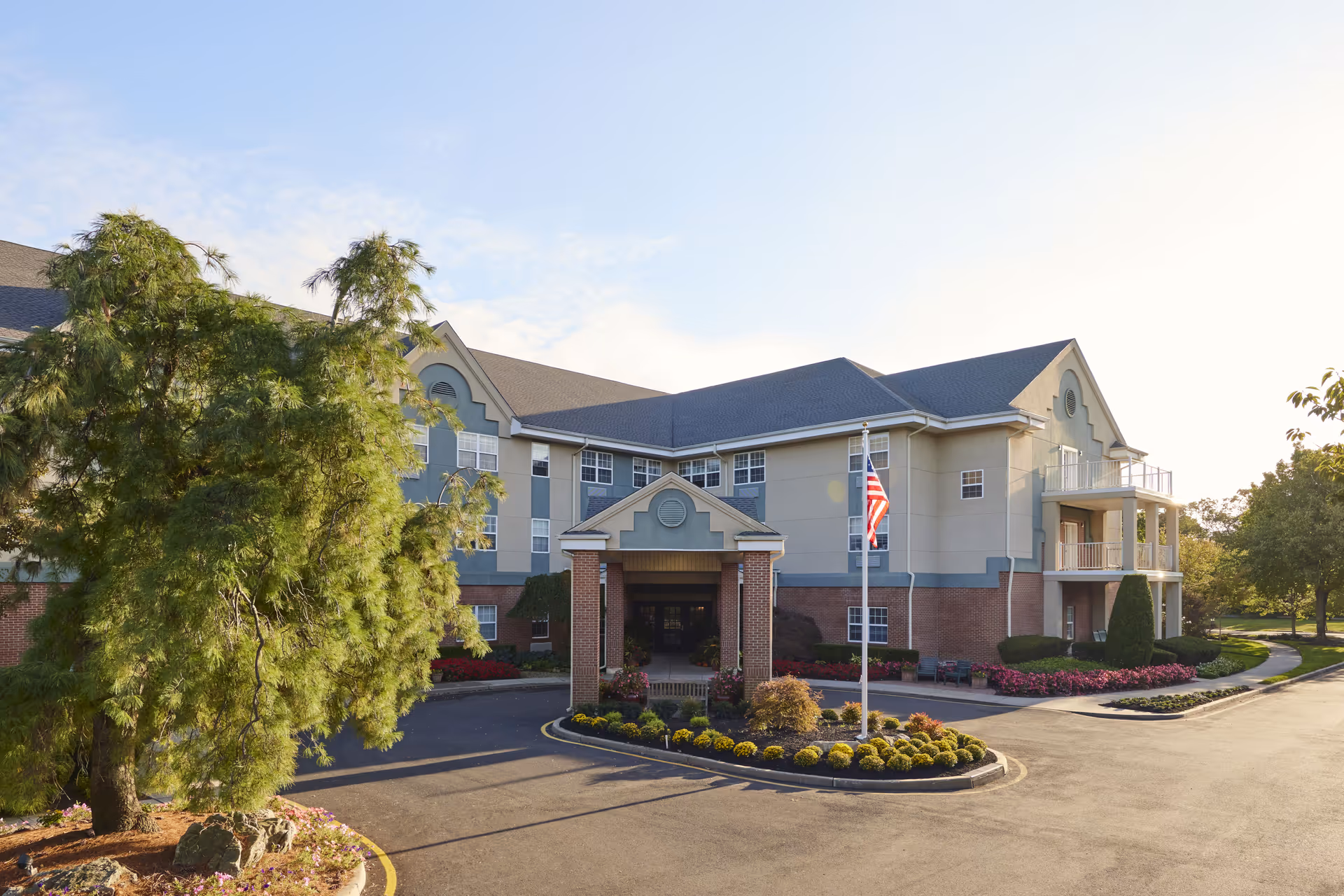 Exterior view of a three-story senior living facility building with a covered entrance supported by brick columns, surrounded by landscaped flower beds and trees, with an American flag on a flagpole in front.