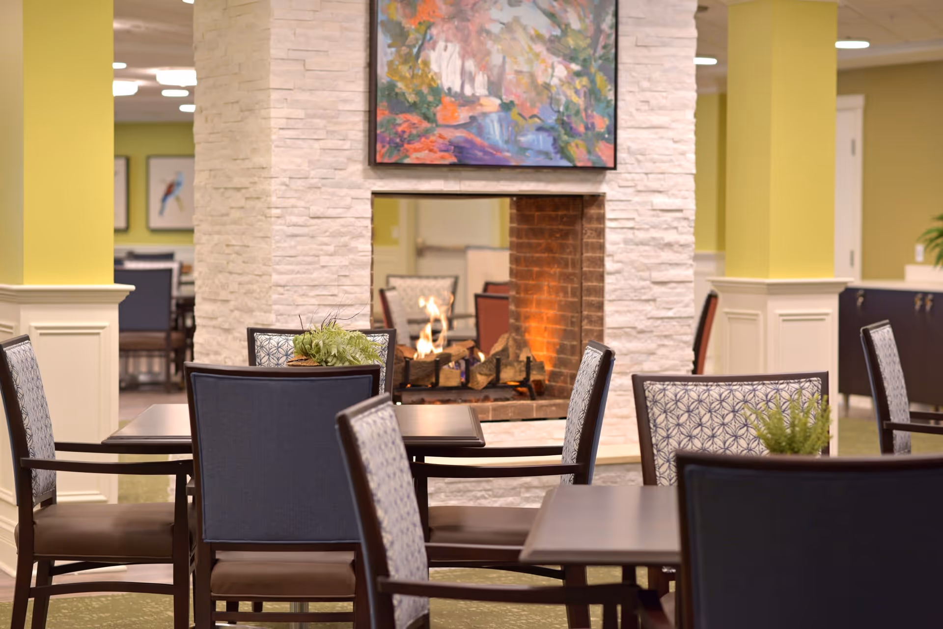 A cozy interior dining area with several tables and chairs arranged around a central stone fireplace with a fire burning. Above the fireplace is a colorful abstract painting. The walls are painted light green with white wainscoting, and there are decorative plants on some tables.