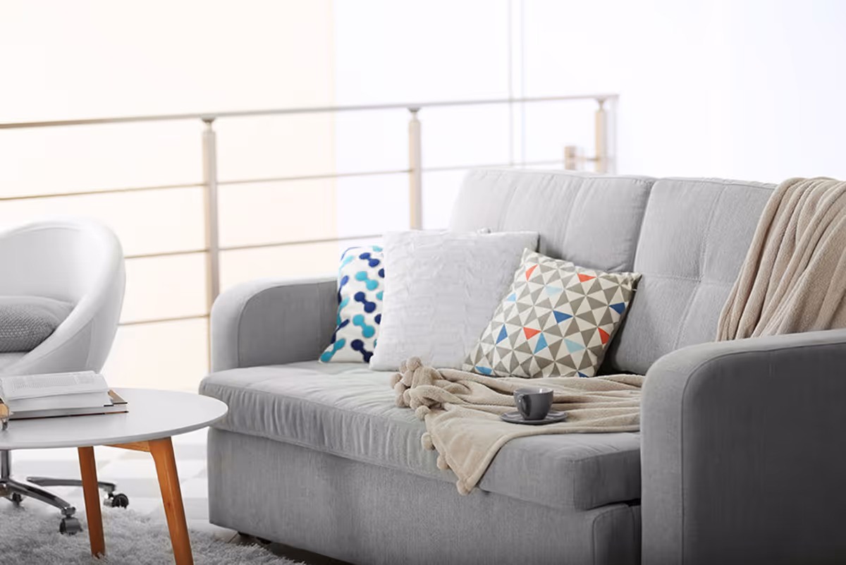 A light gray sofa with patterned throw pillows and a blanket beside a round coffee table and chair in a bright living area.