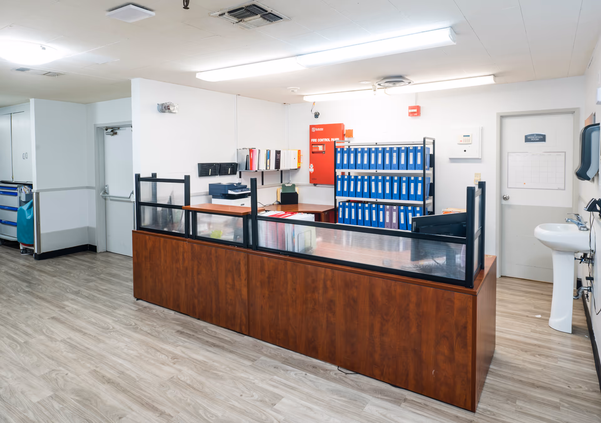 Reception/administrative workstation inside a facility with a long wood counter, shelving filled with blue binders, a wall sink, and overhead lighting.