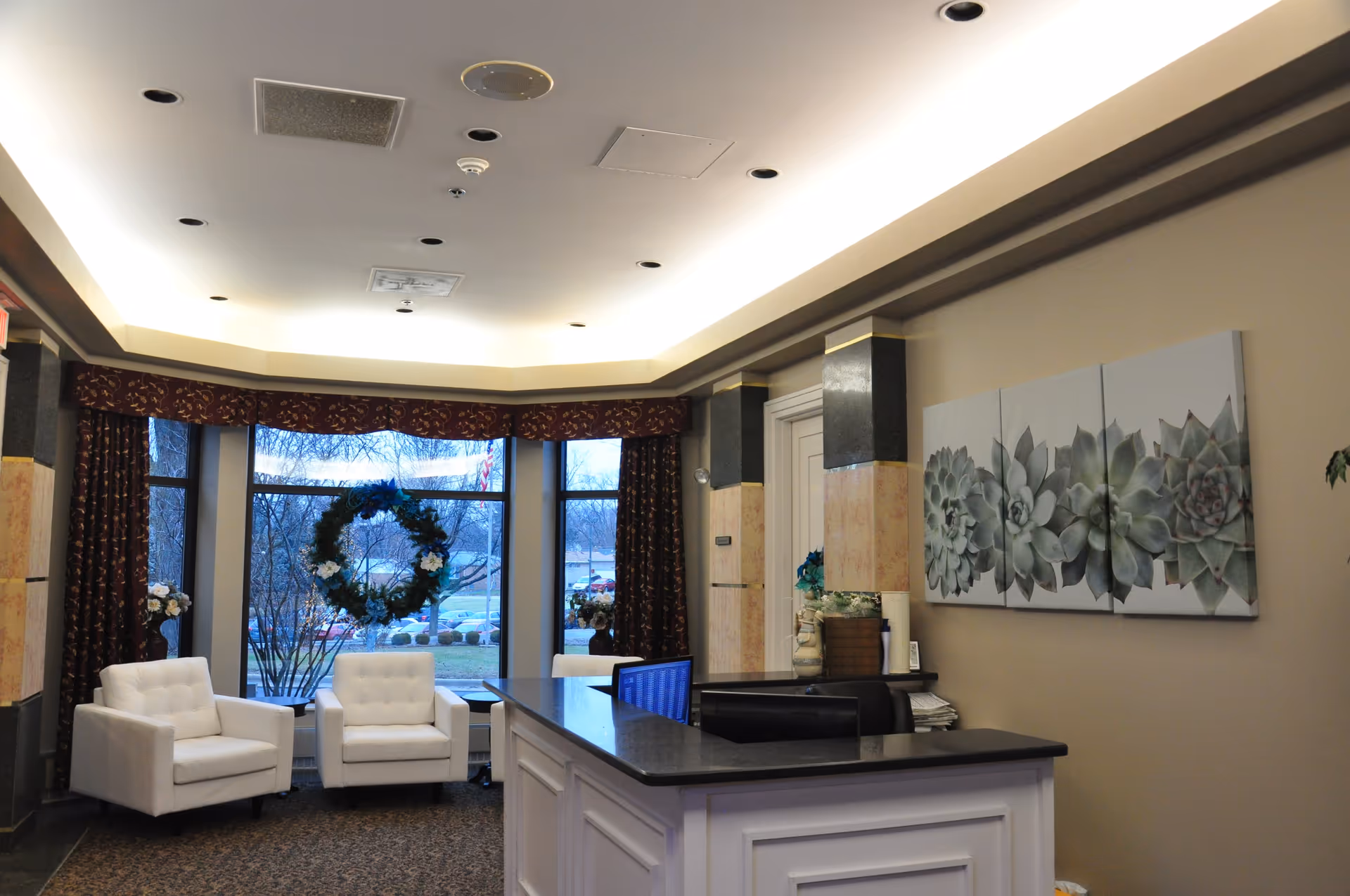 Reception area with a white desk and two computer monitors, two white armchairs near large windows decorated with a green wreath, floral curtains, and a triptych wall art featuring succulent plants.