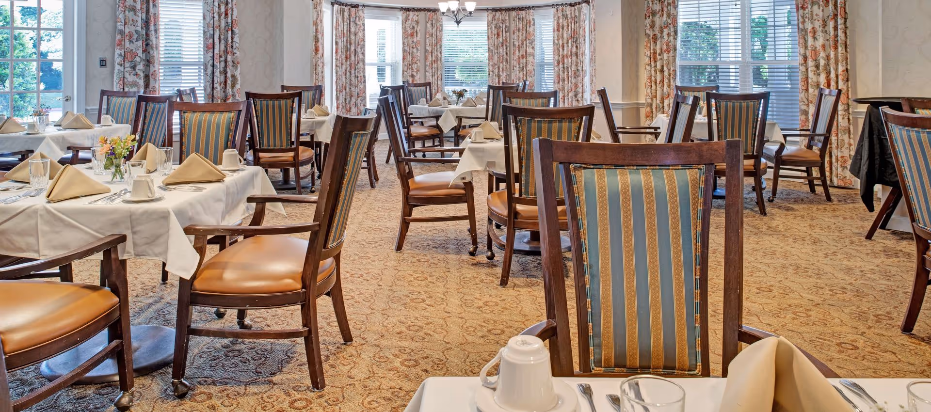 Formal dining room with multiple set tables, striped upholstered wooden chairs, and large windows with floral curtains.