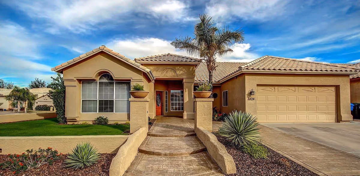 Front exterior view of a single-story house with a tiled roof, a large window, a wooden front door, a two-car garage, and a landscaped yard with palm trees and desert plants under a partly cloudy sky.