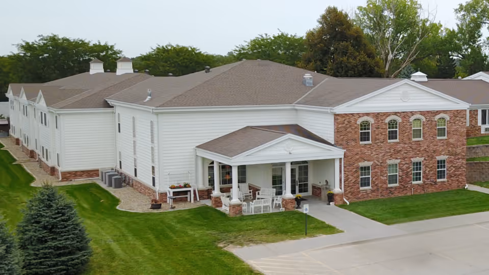 Exterior view of a two-story senior living facility building with white siding and red brick accents. The building has a covered entrance with white columns and outdoor seating. Surrounding the building is a well-maintained lawn with trees in the background and a paved driveway in front.