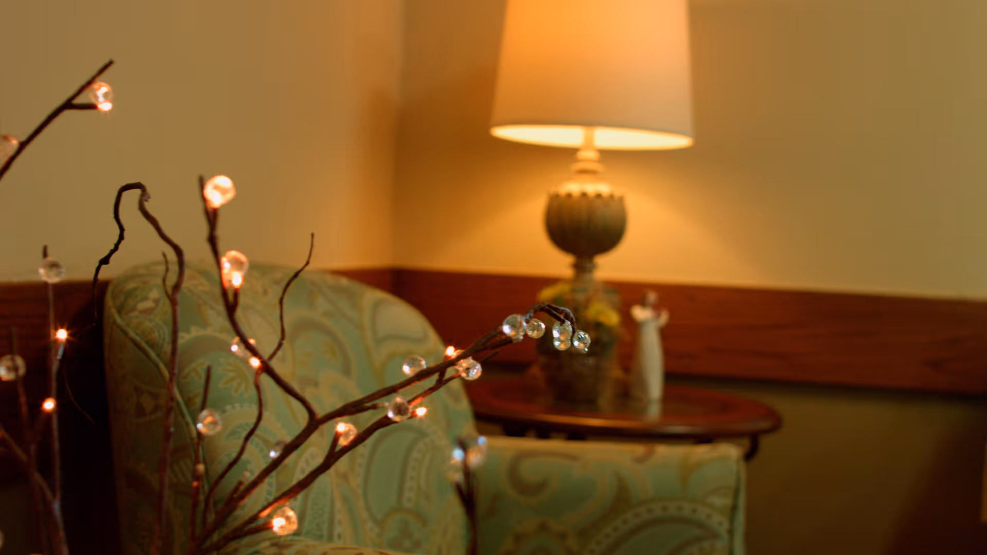 A cozy corner of a room featuring a patterned green armchair, a wooden side table with a decorative lamp, and a small figurine. Twigs with small glowing lights are in the foreground, creating a warm and inviting atmosphere.