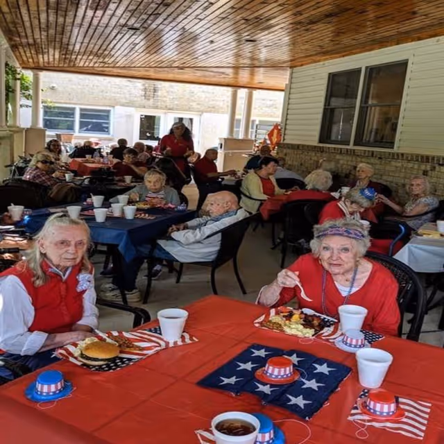 A group of elderly people sitting at tables covered with red, white, and blue decorations, enjoying a meal together under a covered outdoor patio area. The tables have patriotic-themed hats and napkins, and the setting appears festive and social.