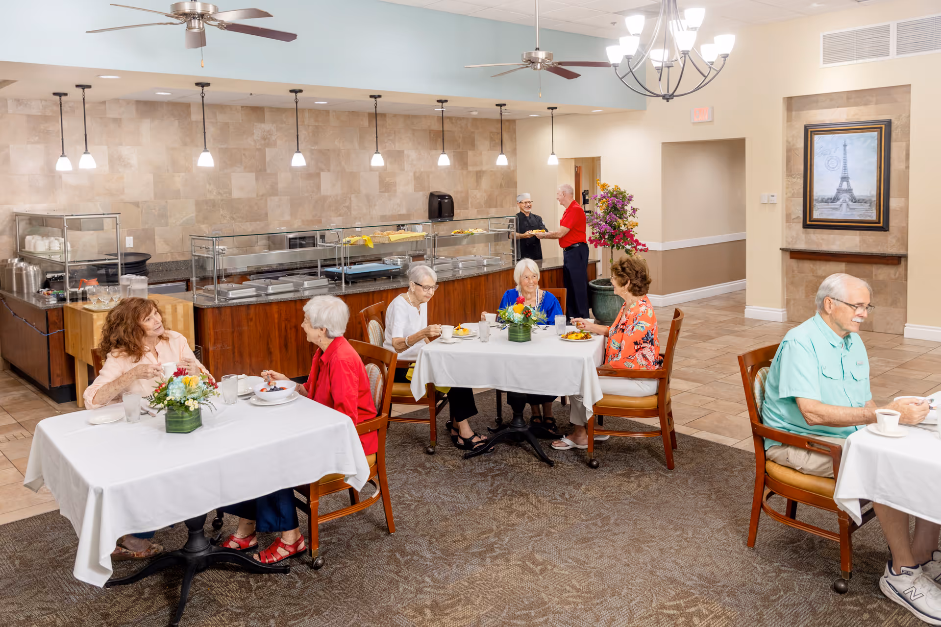 Seniors seated at small tables in a communal dining room with a buffet line and staff serving food.