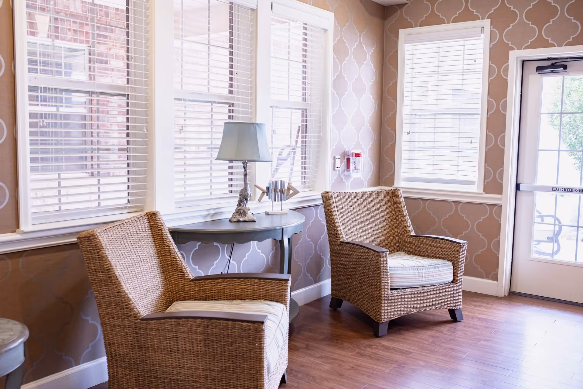 A cozy sitting area with two wicker armchairs featuring striped cushions, positioned near windows with white blinds. A small round table with a decorative lamp and a modern sculpture sits between the chairs. The walls have a patterned wallpaper, and there is a glass door with a 'Push to Exit' sign leading outside.
