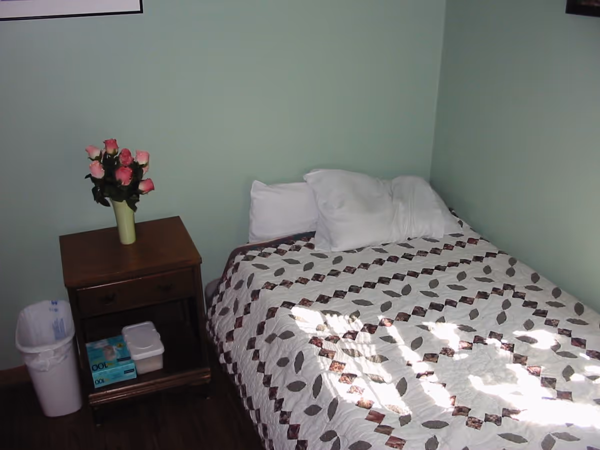 A small bedroom with a bed covered in a patterned quilt and two white pillows. Next to the bed is a wooden nightstand with a vase of pink roses on top, a box of gloves, and a small container on the lower shelf. A white trash bin is placed beside the nightstand. The walls are painted light green.