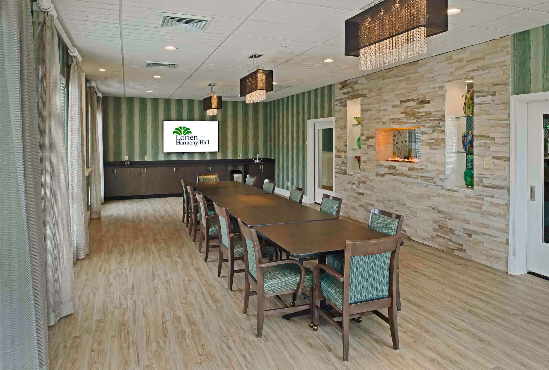 A spacious dining room with a long rectangular table surrounded by chairs with green upholstery. The room features wood flooring, green striped wallpaper, and a stone accent wall with built-in shelves displaying decorative glass items. Modern chandeliers hang from the ceiling, and a flat screen on the far wall displays the Lorien Harmony Hall logo.