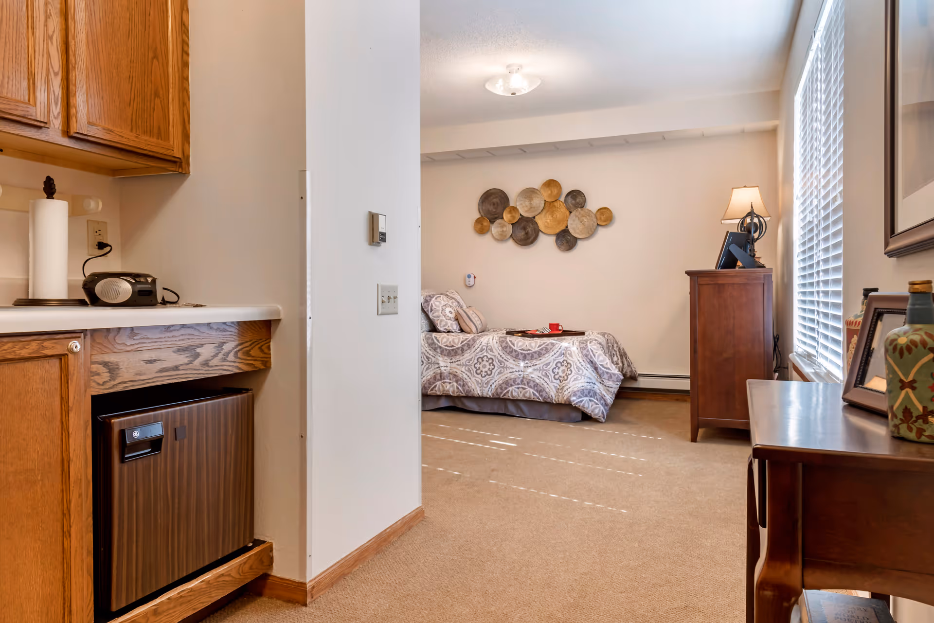 Interior view of a senior living facility room at Brookdale West St. Paul showing a small kitchenette area with wooden cabinets and a mini fridge on the left, and a bedroom area with a single bed covered in patterned bedding, a wooden dresser with a lamp, and a decorative wall hanging. There is a window with blinds on the right side letting in natural light.