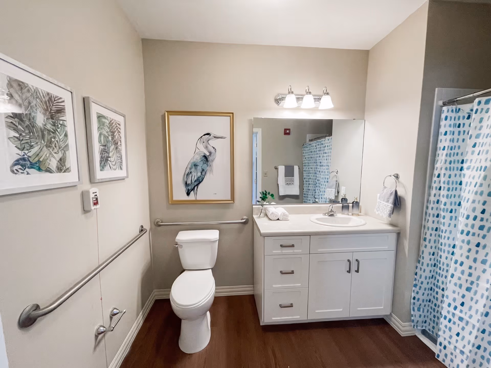 Bright accessible bathroom with a toilet, vanity and sink under a large mirror, a shower with a blue patterned curtain, grab bars and framed artwork.