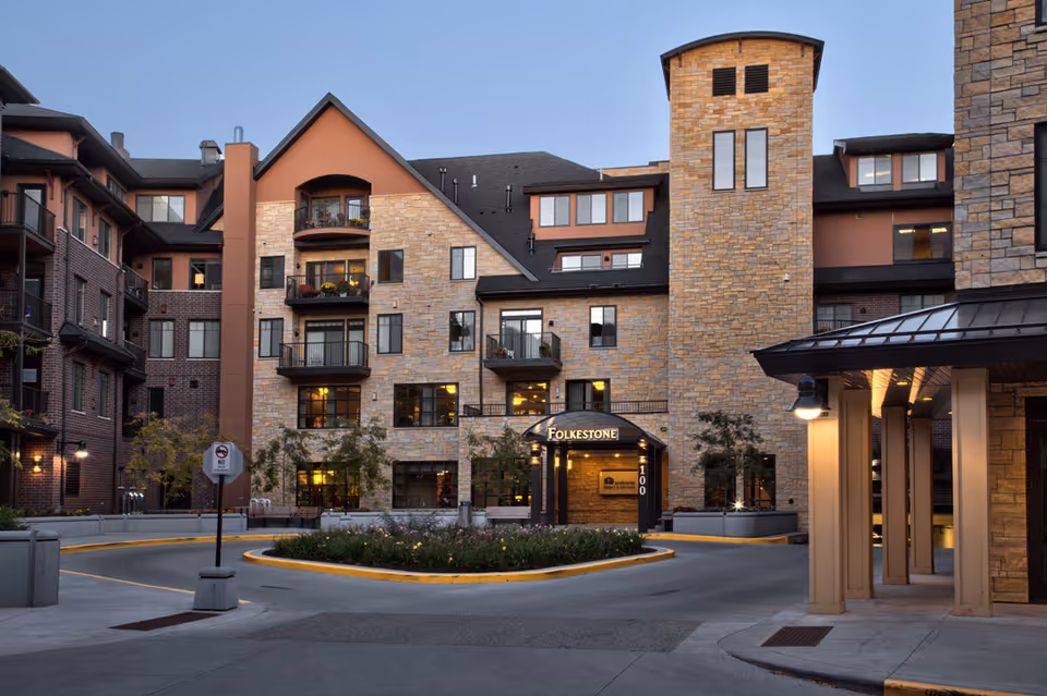 Exterior view of the Folkestone senior living facility building at dusk, showing a multi-story structure with stone and brick facade, balconies, windows, and a covered entrance with the Folkestone sign illuminated.