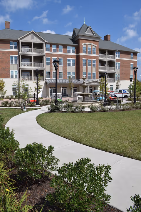 Front view of a multi-story brick senior living building with balconies, a central fountain, a curved walkway, and landscaped lawn under a blue sky.