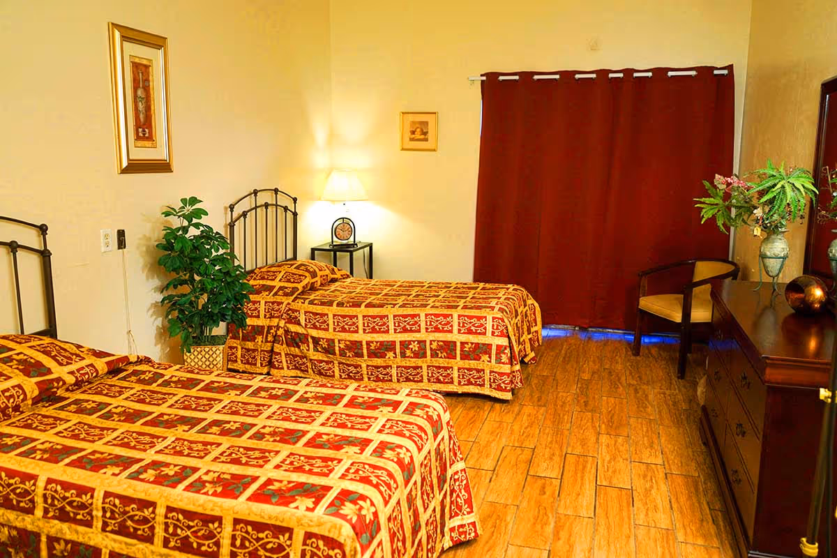 A bedroom with two single beds covered in red and gold patterned bedspreads. There is a small black nightstand with a lamp and clock between the beds. A potted plant is placed beside the nightstand. The room has wooden flooring, a dresser with a mirror on the right side, a chair, and a vase with flowers. Dark red curtains cover the window.