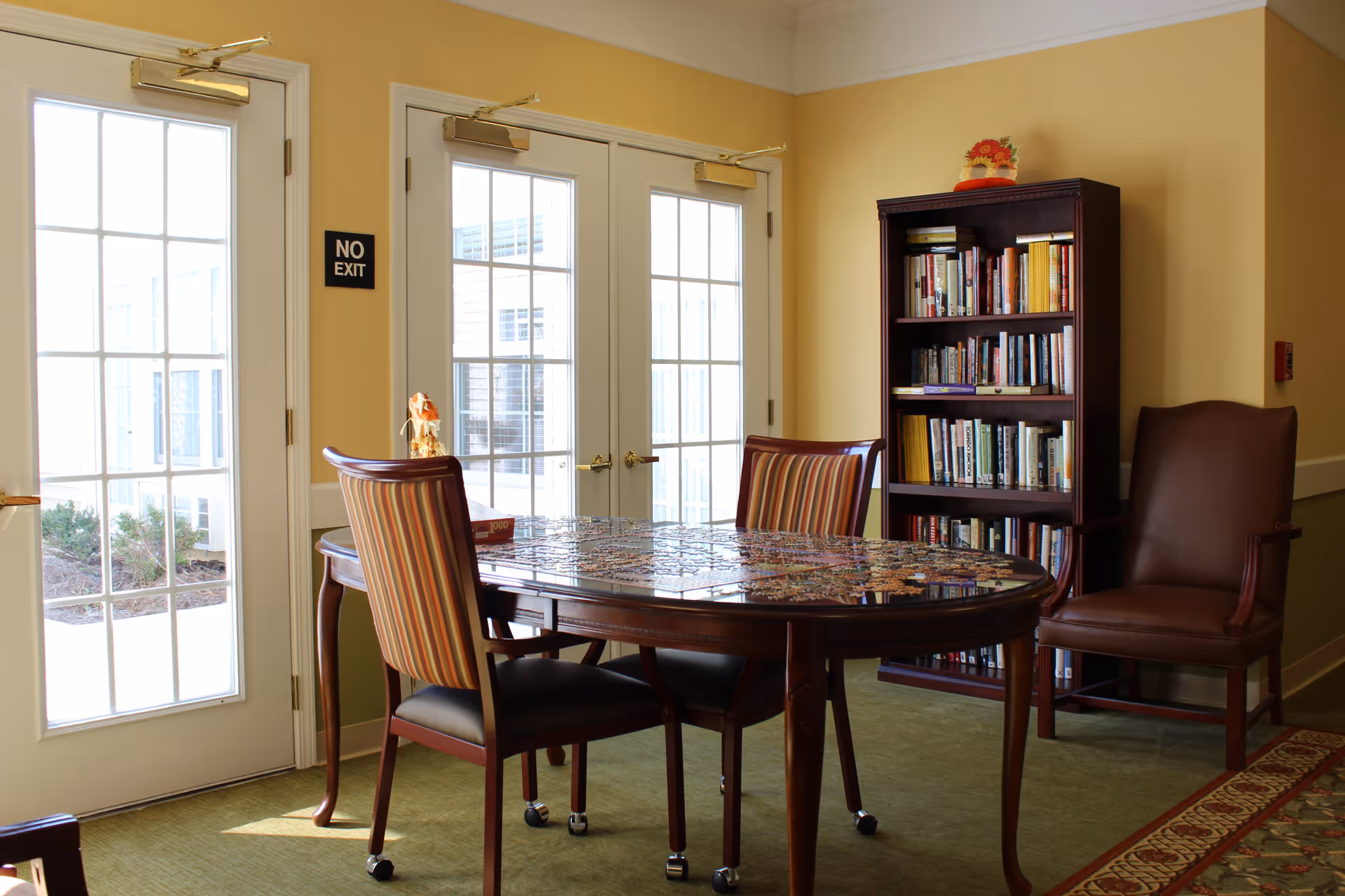 A cozy room with a wooden table in the center covered with a partially completed jigsaw puzzle. Two striped chairs and one brown leather chair surround the table. A tall bookshelf filled with books stands against a yellow wall. French doors with glass panes let in natural light, and a 'NO EXIT' sign is mounted on the wall between the doors.