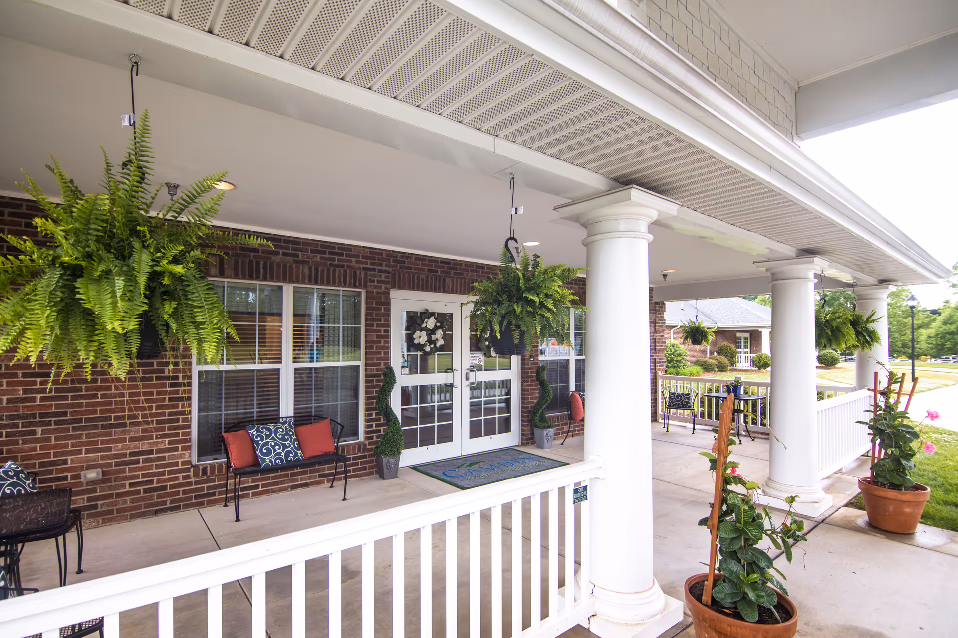 Covered porch area of a senior living facility with white columns, hanging green ferns, potted plants, benches with cushions, and a glass door entrance with decorative wreaths.