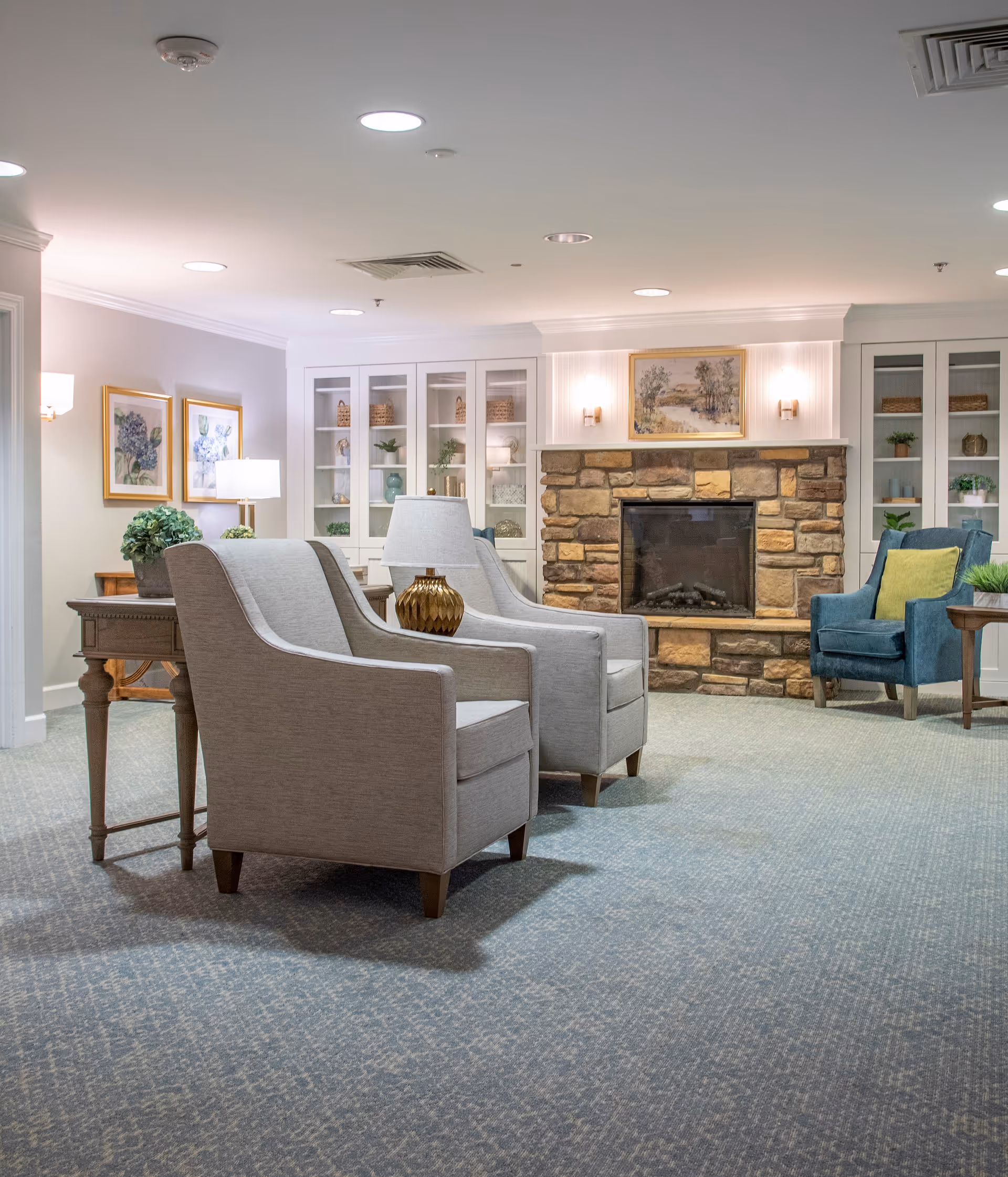 A cozy living room area with two gray armchairs and a blue armchair arranged around a stone fireplace. The room features built-in white cabinets with decorative items, framed artwork on the walls, and soft lighting from wall sconces and table lamps. The carpet is patterned in shades of blue and gray.