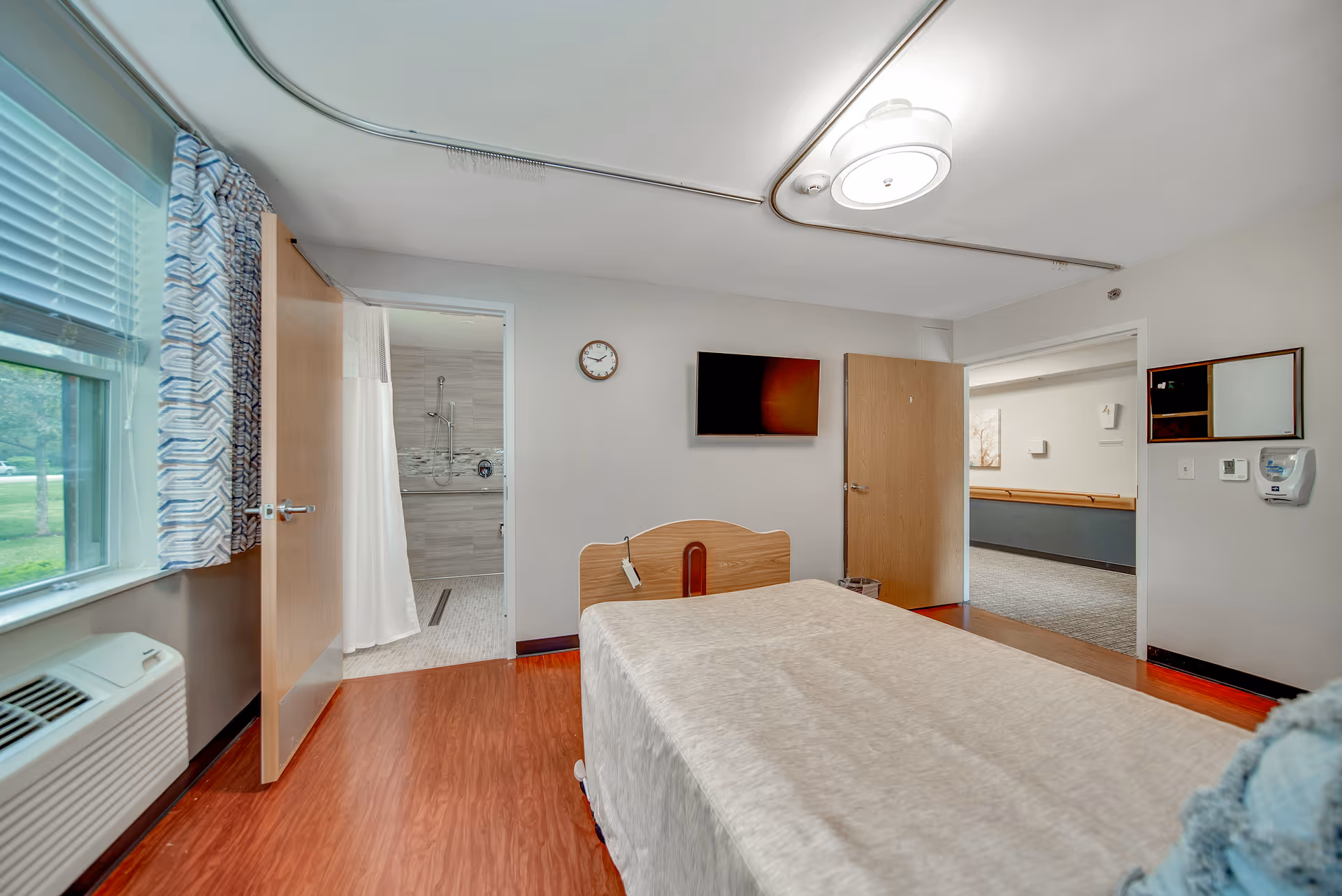A simple, clean bedroom in a healthcare facility with a single bed covered in a beige blanket, a wall-mounted TV, a clock, and two open doors showing a bathroom with a shower and a hallway. There is a window with patterned curtains and an air conditioning unit below it.