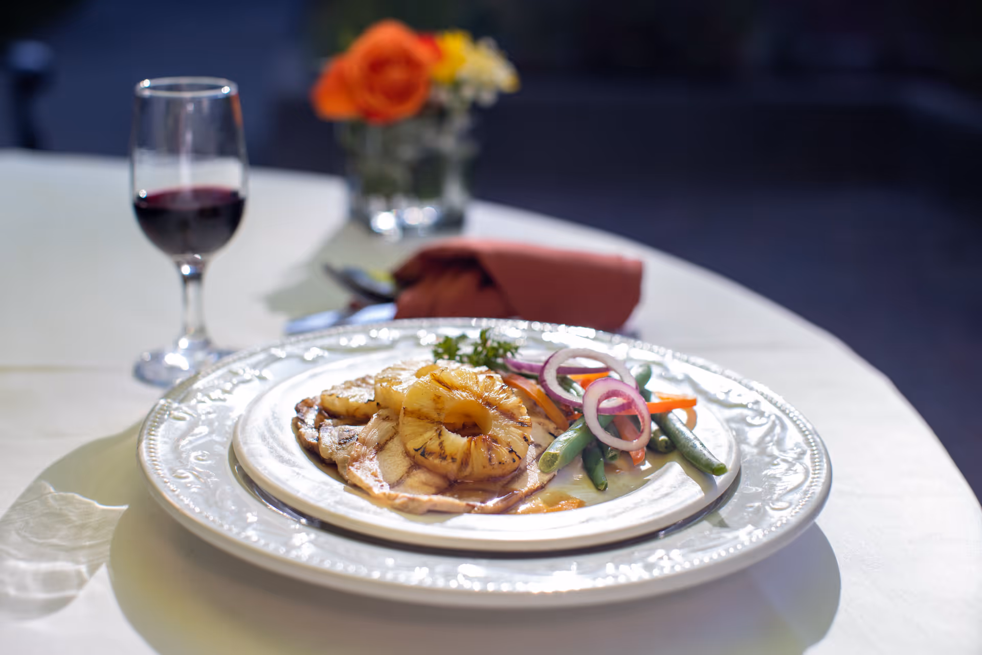 A plated meal featuring grilled pineapple rings on top of a piece of meat, served with green beans, carrot slices, and red onion rings on a white decorative plate. A glass of red wine and a small vase with orange and yellow flowers are in the background on a white tablecloth.