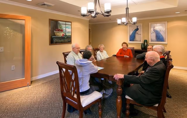A group of elderly residents seated around a large wooden table in a well-lit common dining room.