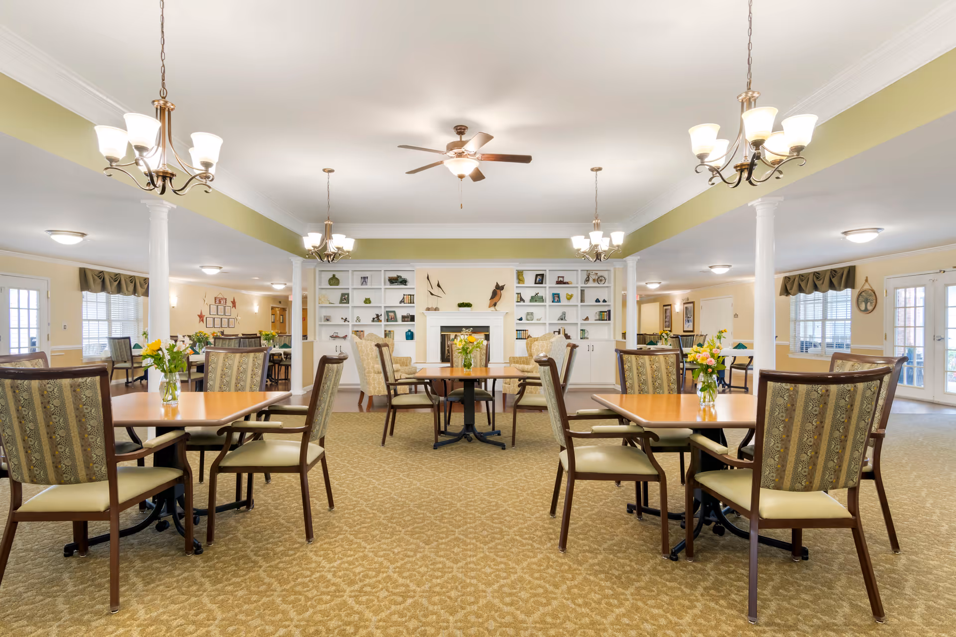 A spacious dining room with multiple wooden tables and cushioned chairs arranged neatly. Each table has a small vase with fresh flowers. The room features beige patterned carpet, white pillars, and a ceiling fan with lights. In the background, there is a white fireplace flanked by built-in shelves decorated with various ornaments and books. The walls are painted in soft yellow and green tones, and there are large windows and glass doors allowing natural light to enter.