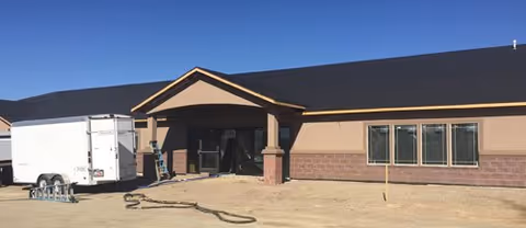 Exterior view of a single-story building with a dark roof and beige walls, featuring a covered entrance supported by columns. A white trailer is parked on the left side on a dirt surface under a clear blue sky.