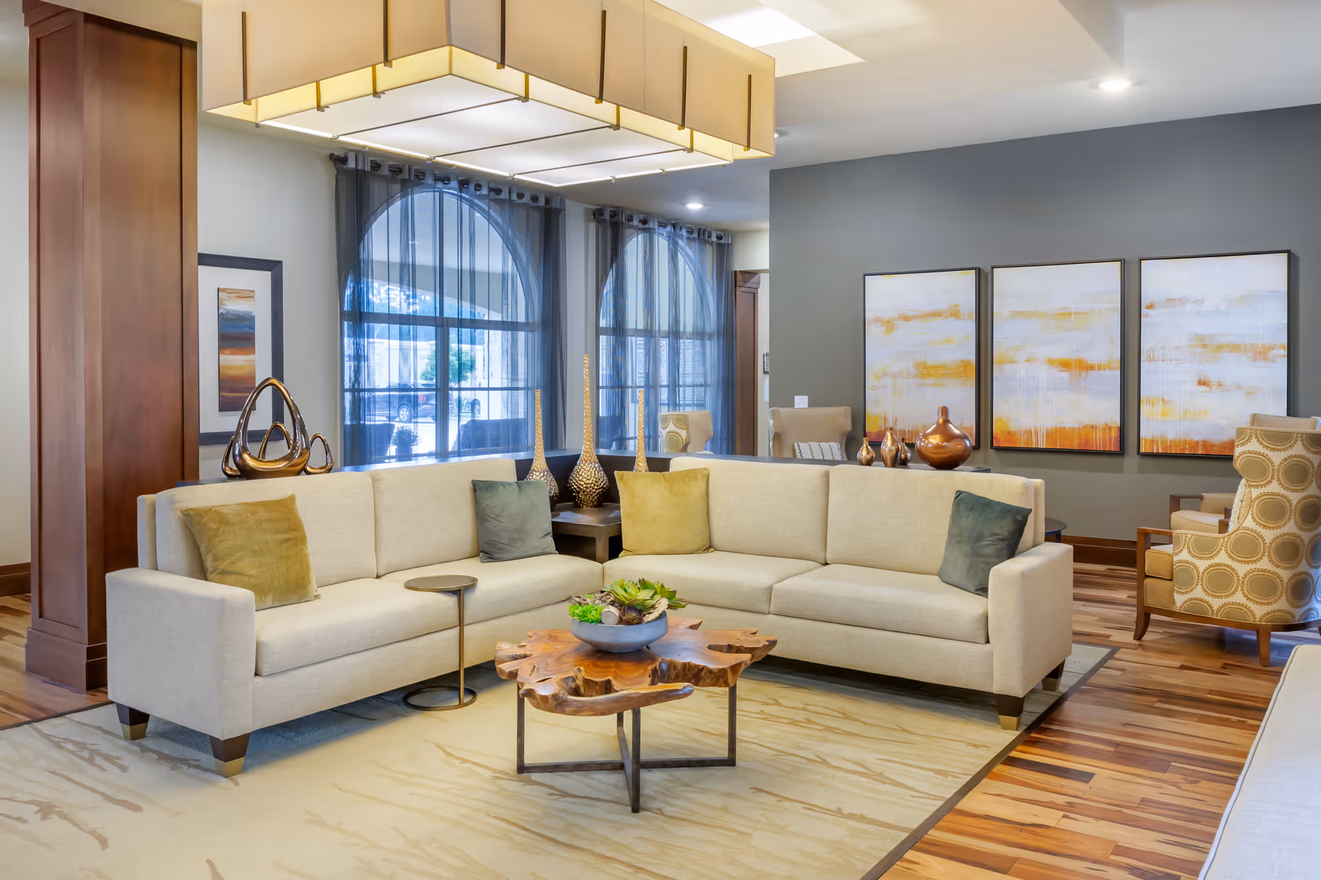 A modern living room with a large beige sectional sofa adorned with green and yellow pillows, a unique wooden coffee table with a plant centerpiece, and two patterned armchairs. The room features large arched windows with sheer curtains, abstract artwork on the wall, and decorative vases on side tables. The floor is a mix of wood and a light-colored area rug, and the ceiling has a large rectangular light fixture.