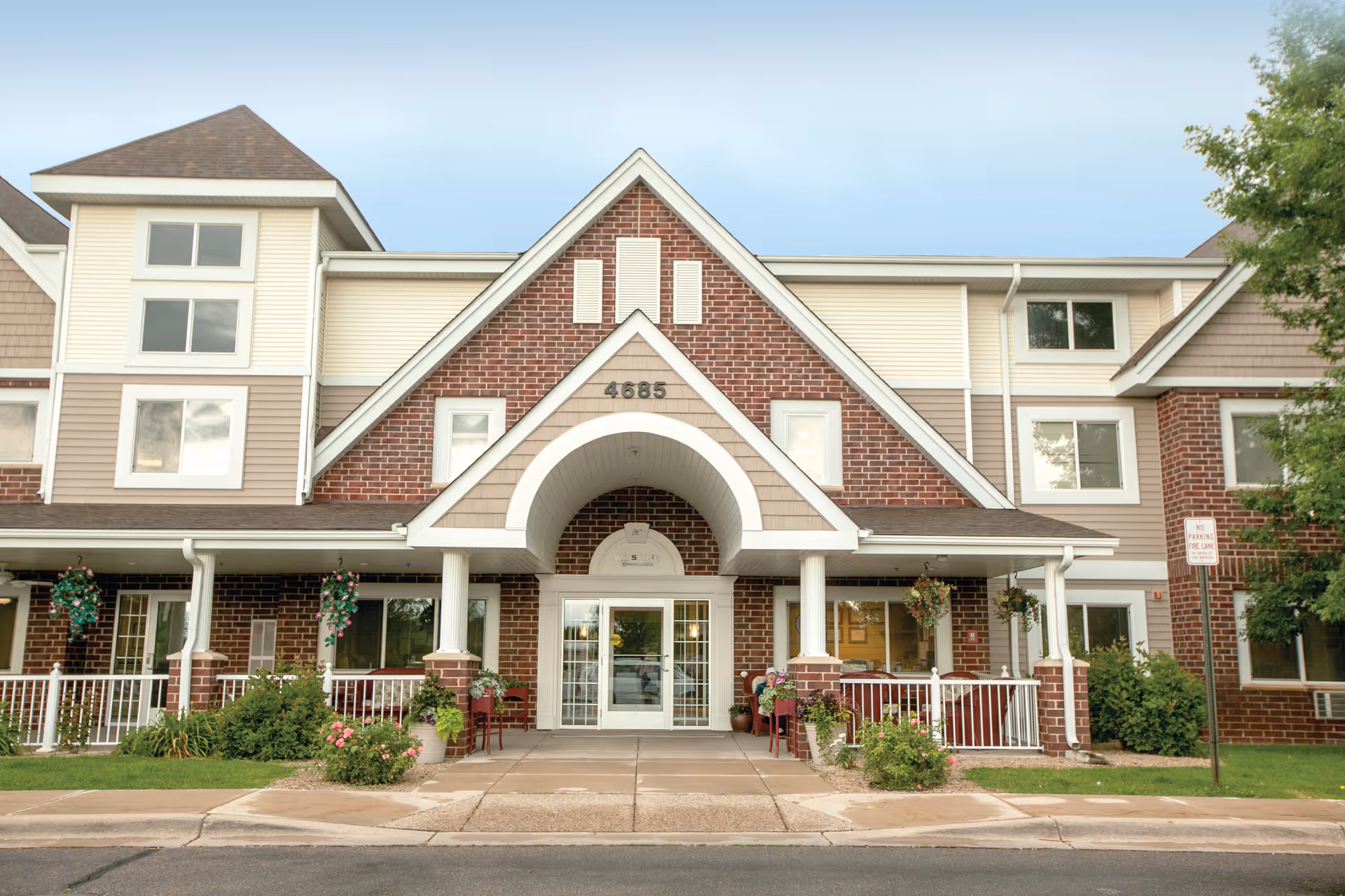 Front exterior view of a senior living facility building with a brick and beige siding facade, a peaked roof entrance with the number 4685 above the door, and a covered porch area with white railings and hanging flower baskets.