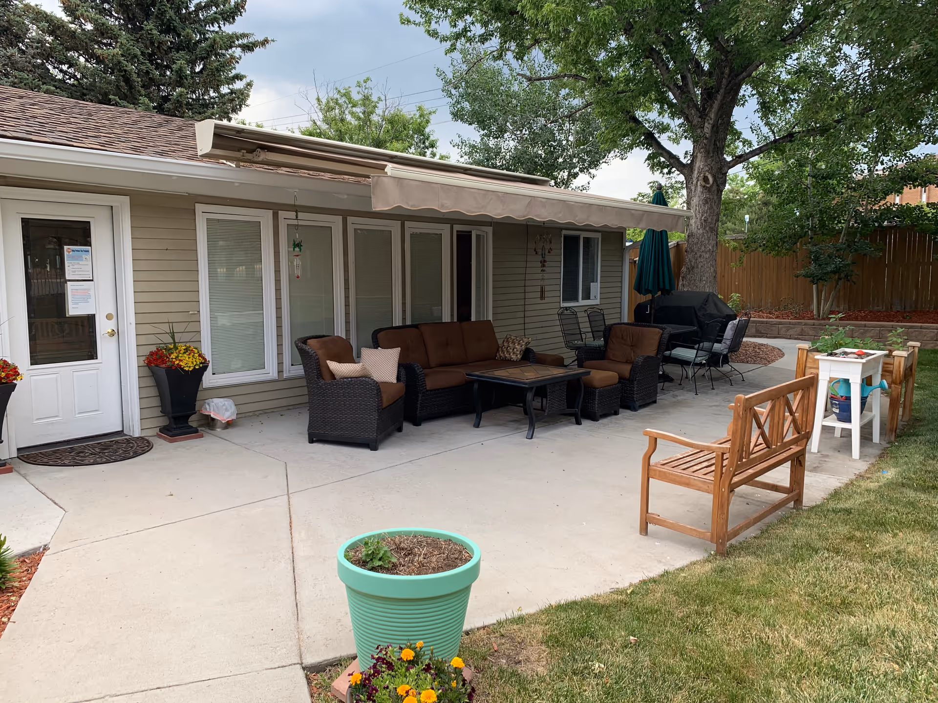 Outdoor patio area of an assisted living facility with cushioned wicker chairs and a sofa around a coffee table, a wooden bench, a small white table with gardening supplies, potted plants, a barbecue grill, and trees in the background.