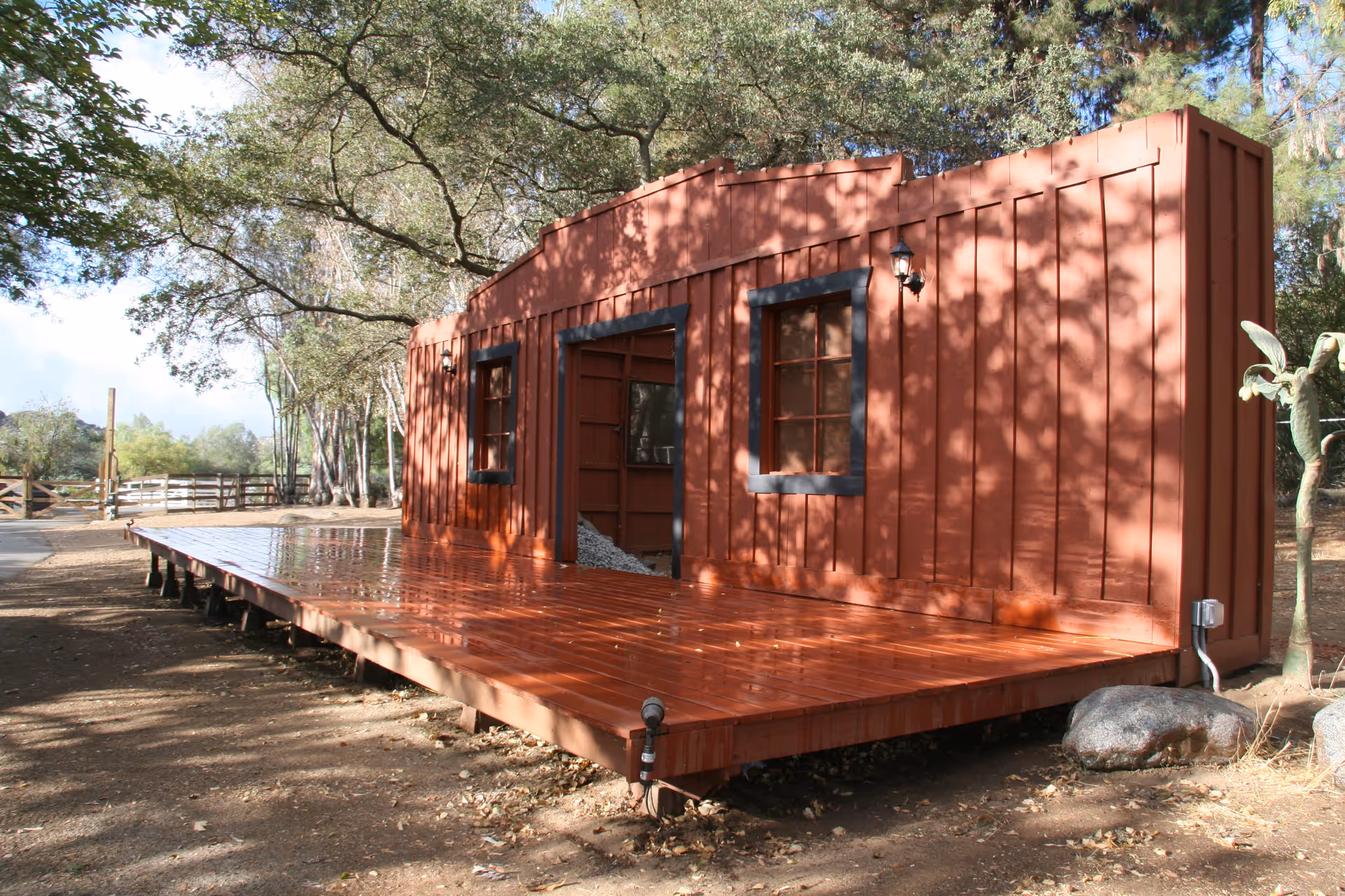 A rustic wooden stage set outdoors resembling the facade of an old western building with two windows and a central doorway, surrounded by trees and natural landscape.