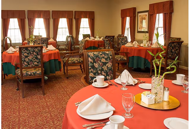 A dining room with round tables covered in red and green tablecloths, set with white napkins, cups, glasses, and silverware. The chairs have floral upholstery, and there are large windows with brown curtains letting in natural light. A decorative plant centerpiece is on the table in the foreground.