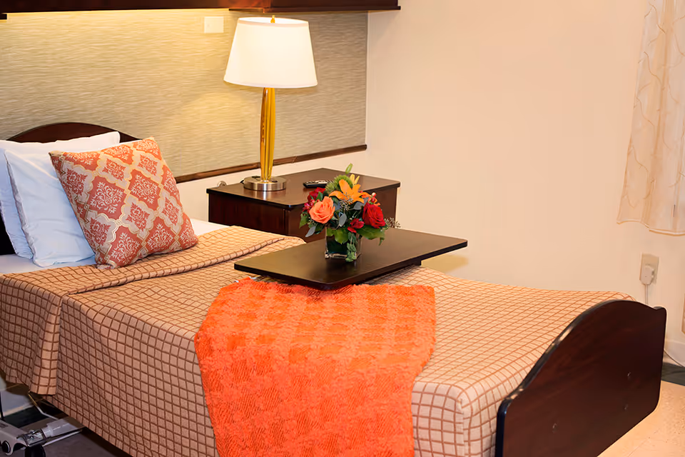 A neatly made single bed with a patterned beige bedspread and an orange throw blanket. There is a decorative pillow with a red and beige pattern, a small wooden bedside table with a lamp, and a small tray holding a vase with colorful flowers. The room has light-colored walls and a window with sheer curtains.