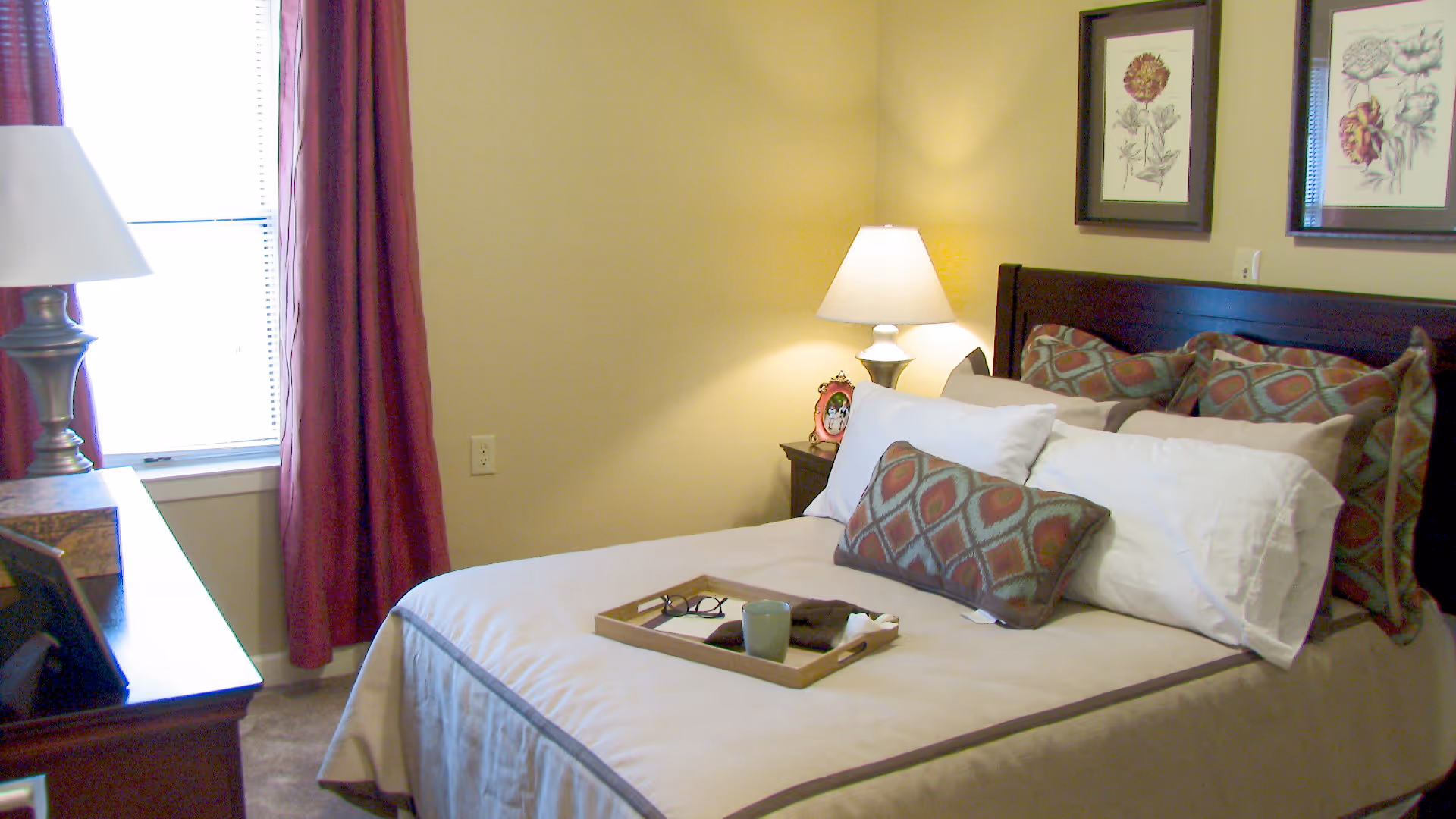 A neatly made bedroom featuring a bed with patterned pillows, a nightstand lamp, a desk, and a window with red curtains.