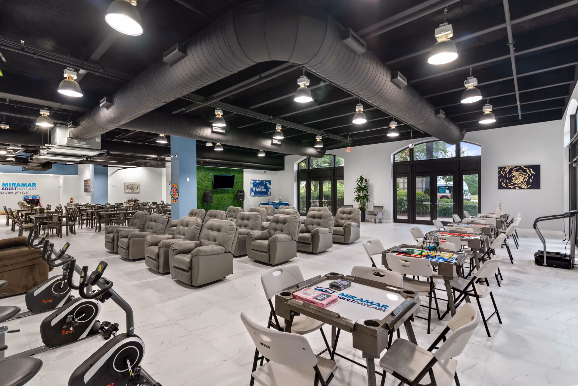 Interior view of Miramar Adult Day Care facility showing a spacious room with multiple rows of comfortable reclining chairs, exercise bikes, and tables with chairs arranged for games or activities. The room has a modern industrial ceiling with exposed ducts and large windows letting in natural light. There is a TV mounted on a green wall and a banner that says Happy Birthday.