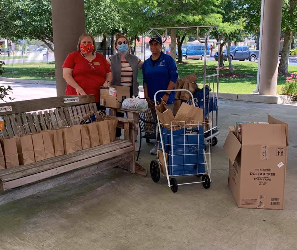 Three women wearing face masks stand under a covered outdoor area next to a bench filled with brown paper bags. There are carts and boxes also filled with bags and supplies. Trees, parked cars, and a grassy area are visible in the background.