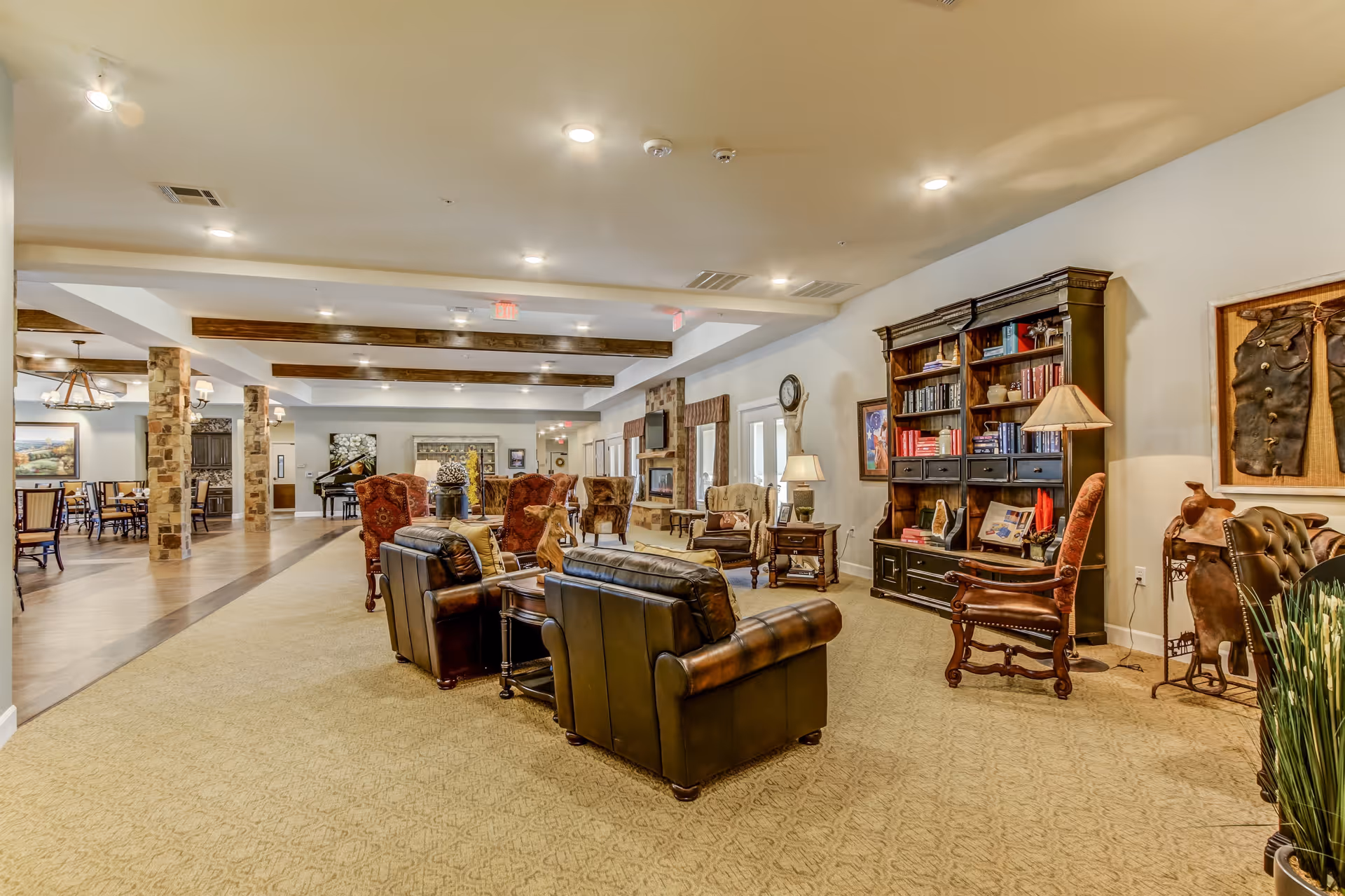 A spacious and well-lit living room area in an assisted living facility featuring leather armchairs, wooden chairs, a large wooden bookshelf filled with books and decorative items, a side table with a lamp, and various wall decorations. The room has carpeted flooring and wooden beams on the ceiling, with a dining area visible in the background.