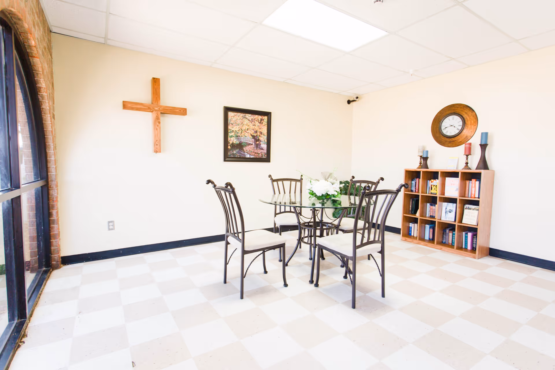 A bright room with a checkered tile floor featuring a glass-top table surrounded by four metal chairs with cushions. On the wall, there is a wooden cross and a framed picture of a tree in autumn. A wooden bookshelf with books, candles, and a round wall clock is also visible. Large windows with brick framing allow natural light into the room.