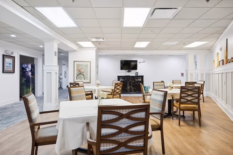 A bright and clean dining room in a senior living facility with several tables covered in white tablecloths and surrounded by wooden chairs with cushioned seats. The room features light wood flooring, white walls with wainscoting, and a flat-screen TV mounted on the far wall above a dark wooden cabinet. There are framed pictures on the walls and small flower arrangements on the tables.
