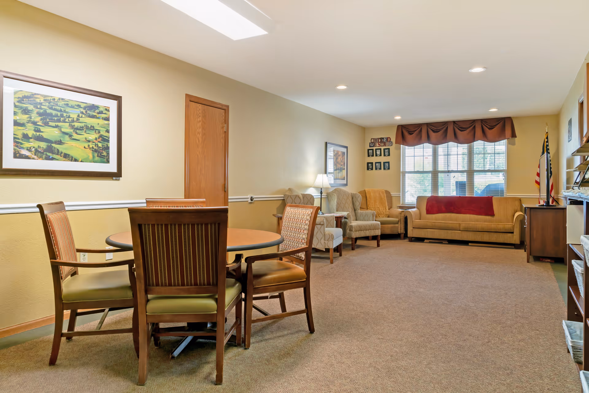 A cozy living room area with a round wooden table surrounded by four chairs in the foreground. In the background, there are two patterned armchairs, a beige sofa with a red throw blanket, a floor lamp, and a large window with brown valances letting in natural light. The walls are painted beige with white trim, and there is a framed landscape painting on the left wall and an American flag near the window.