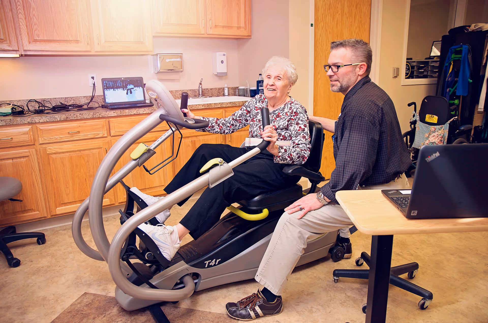 An elderly woman is seated on a recumbent exercise bike in a room with wooden cabinets and a countertop. A man, possibly a caregiver or therapist, is sitting beside her, providing support and encouragement. A laptop is open on a nearby table, and another laptop is on the countertop in the background.