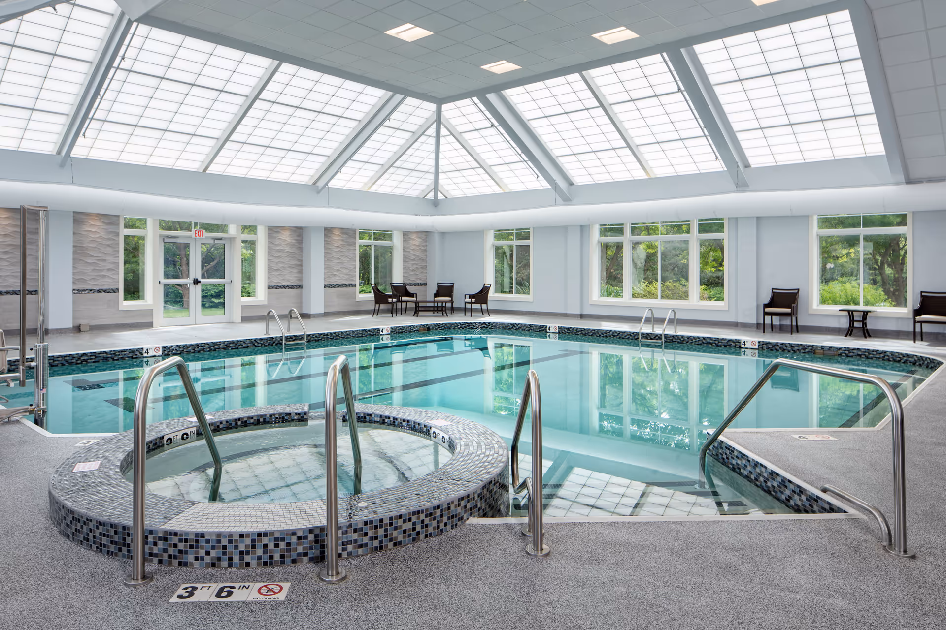 Indoor swimming pool area with a hot tub in the foreground, surrounded by tiled flooring and metal handrails. The pool room has large windows showing greenery outside and a high ceiling with large skylights allowing natural light to fill the space. Several chairs and small tables are placed along the walls.