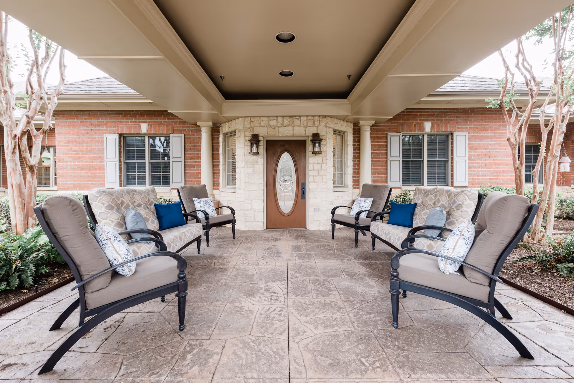 Covered outdoor seating area with cushioned chairs and loveseats arranged facing each other on a stamped concrete floor, flanked by brick walls with windows and a central wooden door with glass panel, surrounded by stone accents and two wall-mounted lanterns.