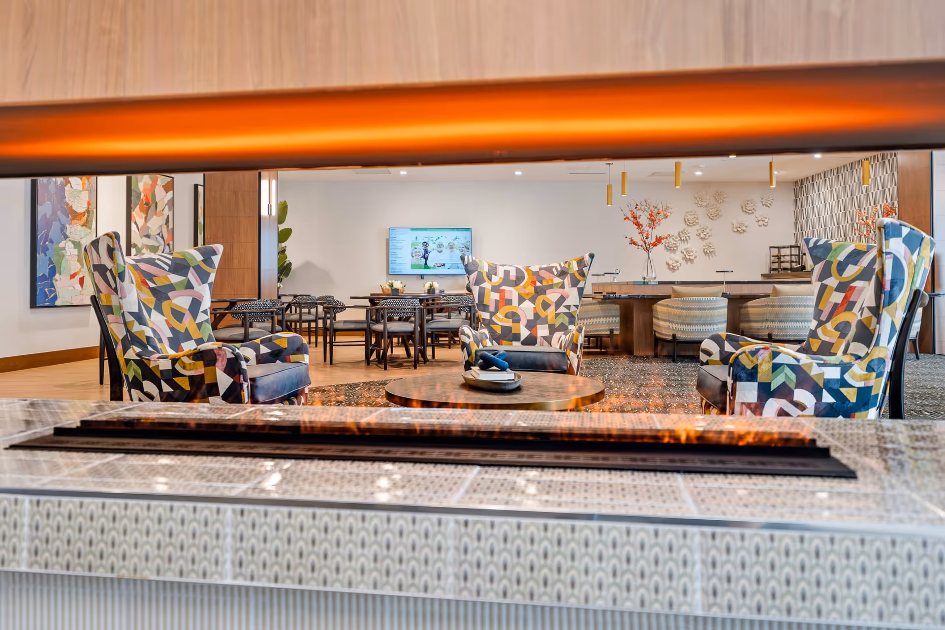 A modern and cozy common area with three colorful patterned armchairs arranged around a round wooden coffee table. In the background, there are dining tables and chairs, a wall-mounted TV, decorative wall art, and a vase with orange flowers. The image is taken from behind a decorative fireplace with a warm flame visible.