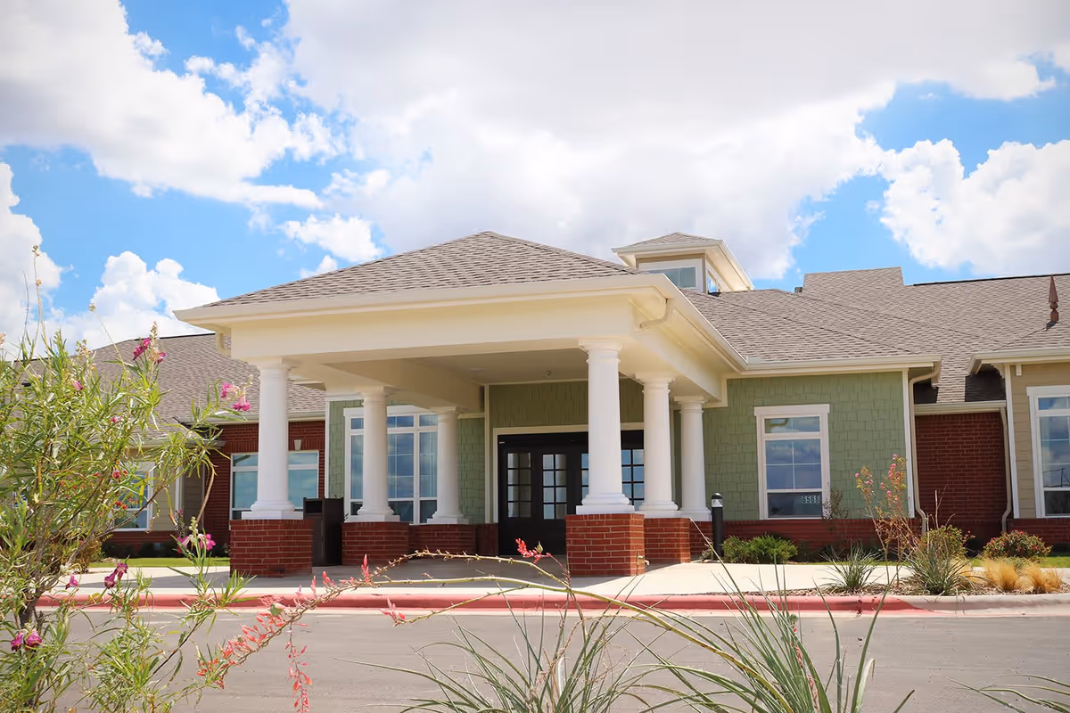 Exterior view of a single-story senior living facility building with a covered entrance supported by white columns, green siding, and brick accents under a partly cloudy blue sky.