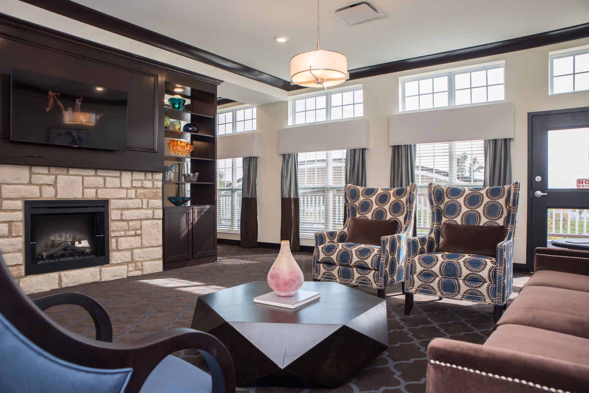 A bright and cozy living room with large windows letting in natural light. The room features a stone fireplace with a mounted flat-screen TV above it, built-in dark wood shelves with decorative glass bowls, two patterned armchairs with brown cushions, a brown sofa, and a geometric dark wood coffee table with a pink vase and a book on top. The walls are light-colored, and the ceiling has a modern light fixture.