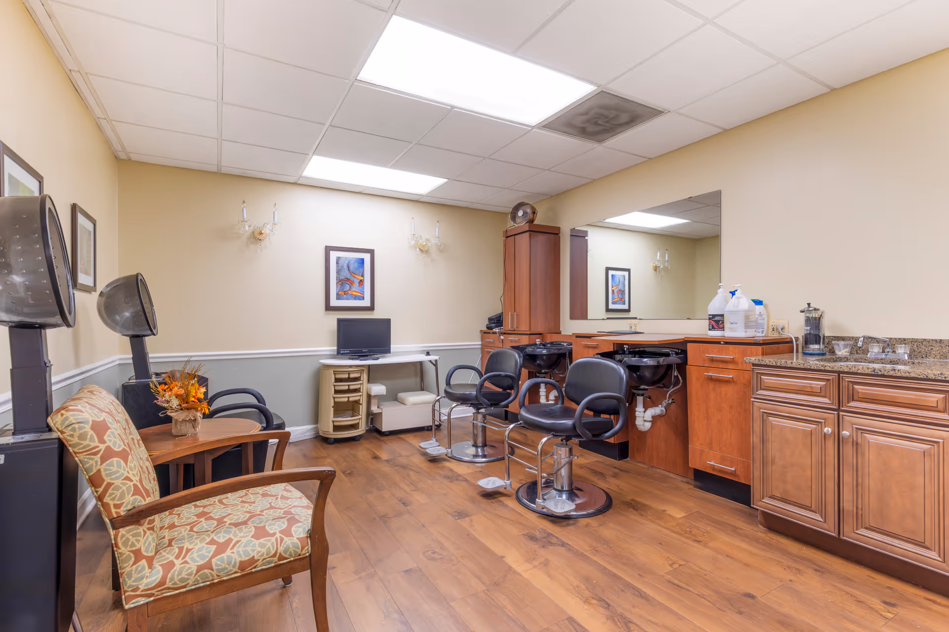 Interior view of a salon area in a senior living facility with two black salon chairs in front of two black hair washing sinks, wooden cabinets, a large mirror, two hair dryers, a patterned armchair, a small table with a flower arrangement, a small TV on a white table, and framed artwork on beige walls.