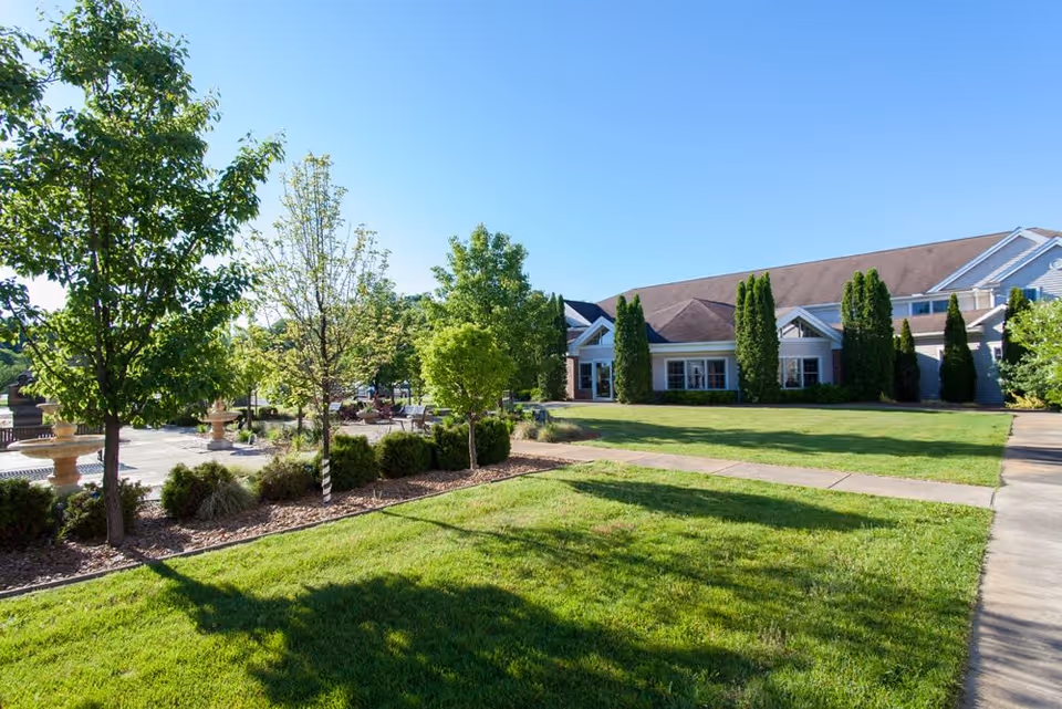 A sunny outdoor view of a senior living facility with a well-maintained lawn, several trees, shrubs, and a paved walkway. The building in the background has a sloped roof and multiple windows, surrounded by tall evergreen trees.
